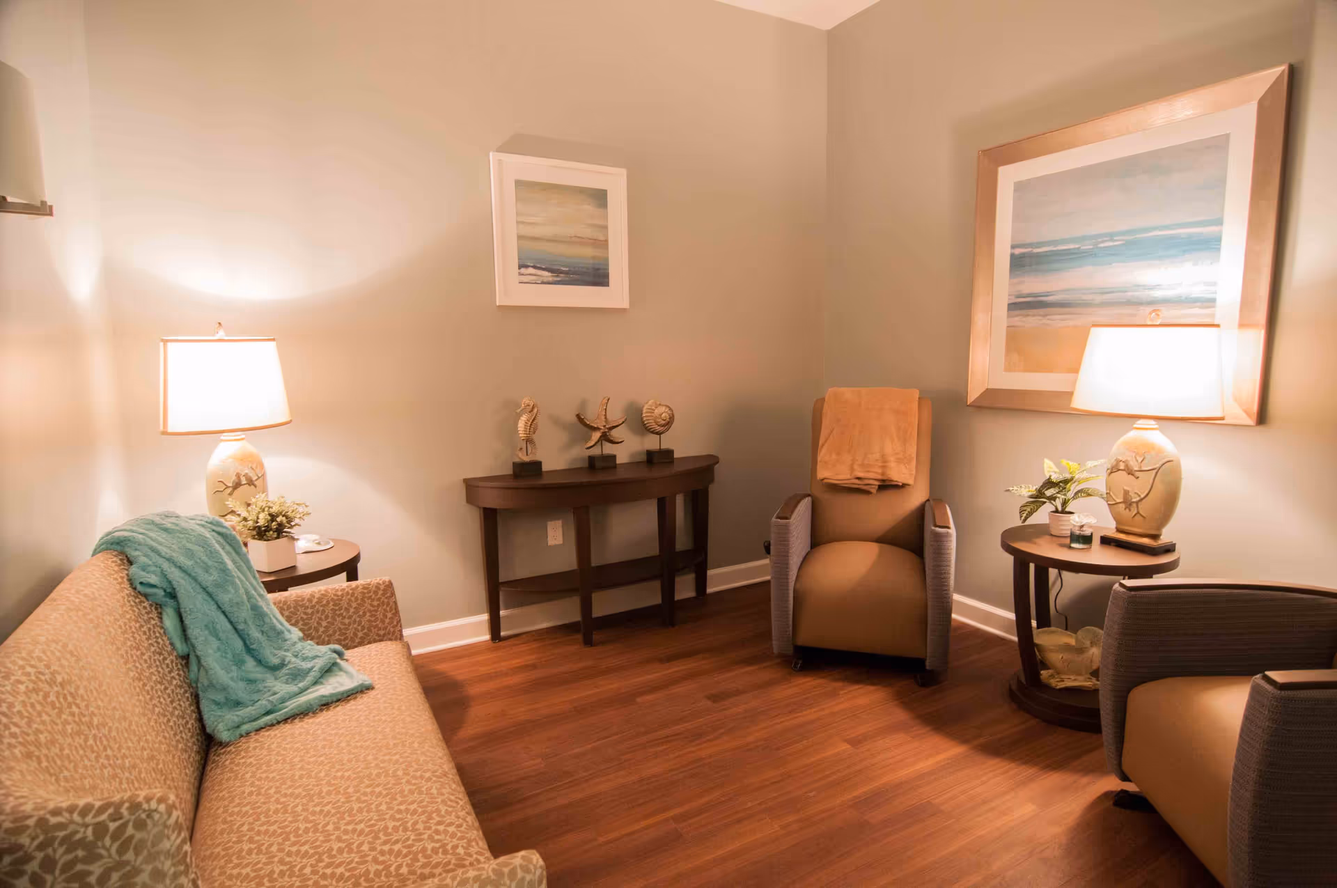A cozy living room area with a patterned beige sofa draped with a teal blanket, two beige armchairs, two side tables each with a lamp and small plants, a wooden console table with three sea-themed sculptures, and two framed paintings on the walls. The room has warm lighting and wooden flooring.