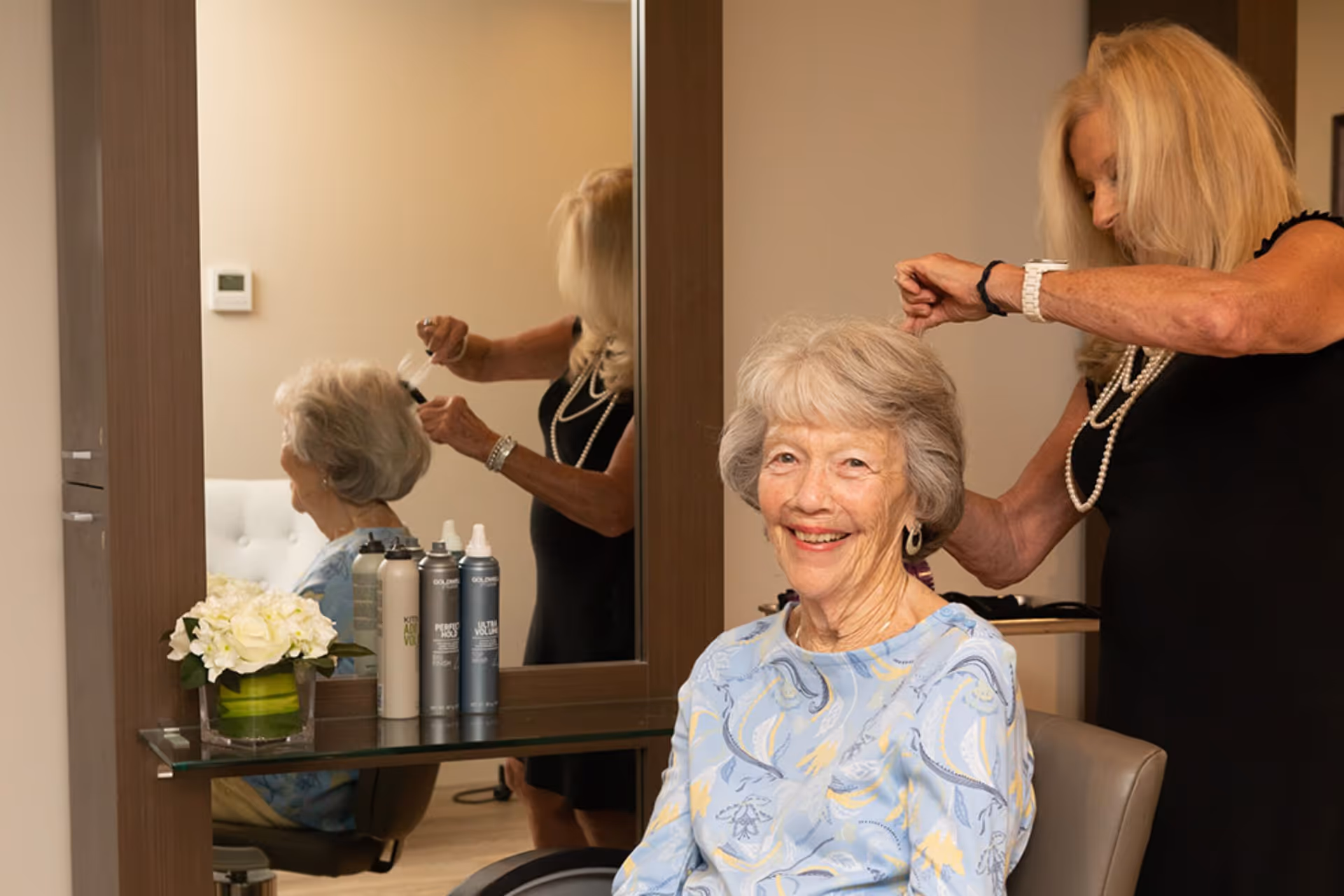 An elderly woman with gray hair and a light blue patterned top is sitting in a chair smiling while a hairstylist with long blonde hair styles her hair in a salon setting. There is a large mirror in front of them reflecting the scene, and several hair product bottles and a vase with white flowers are on a glass shelf beneath the mirror.