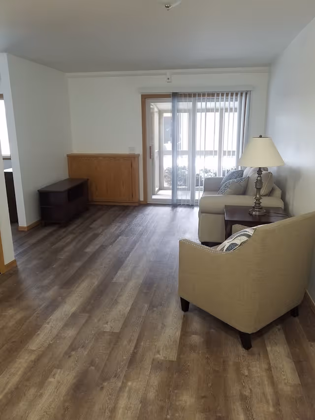 A small living room with wood-look flooring, a beige armchair and sofa next to a side table and lamp, and sliding glass doors with vertical blinds.