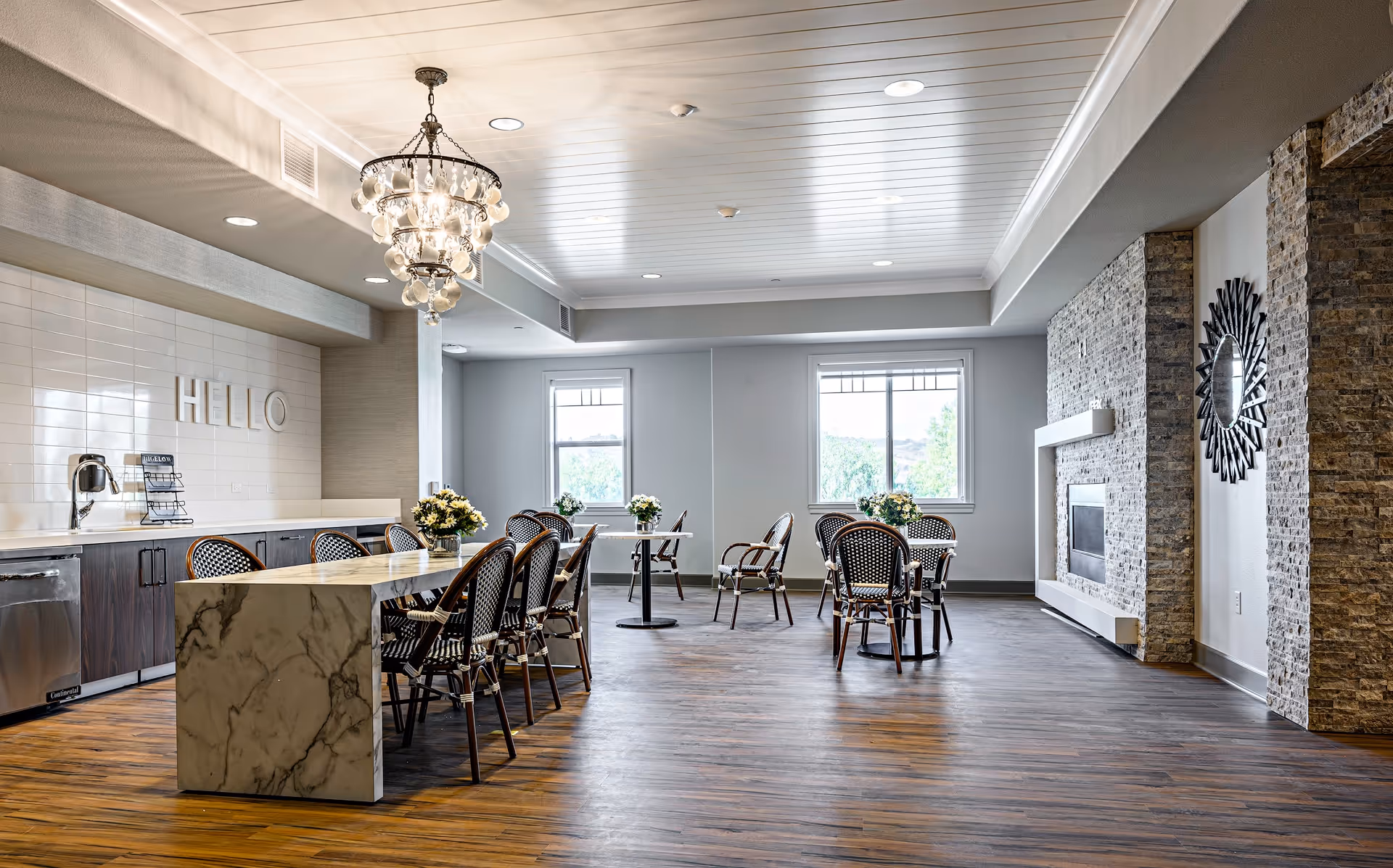 Bright communal dining and lounge area with a marble island, tables and chairs, a chandelier, and a fireplace.
