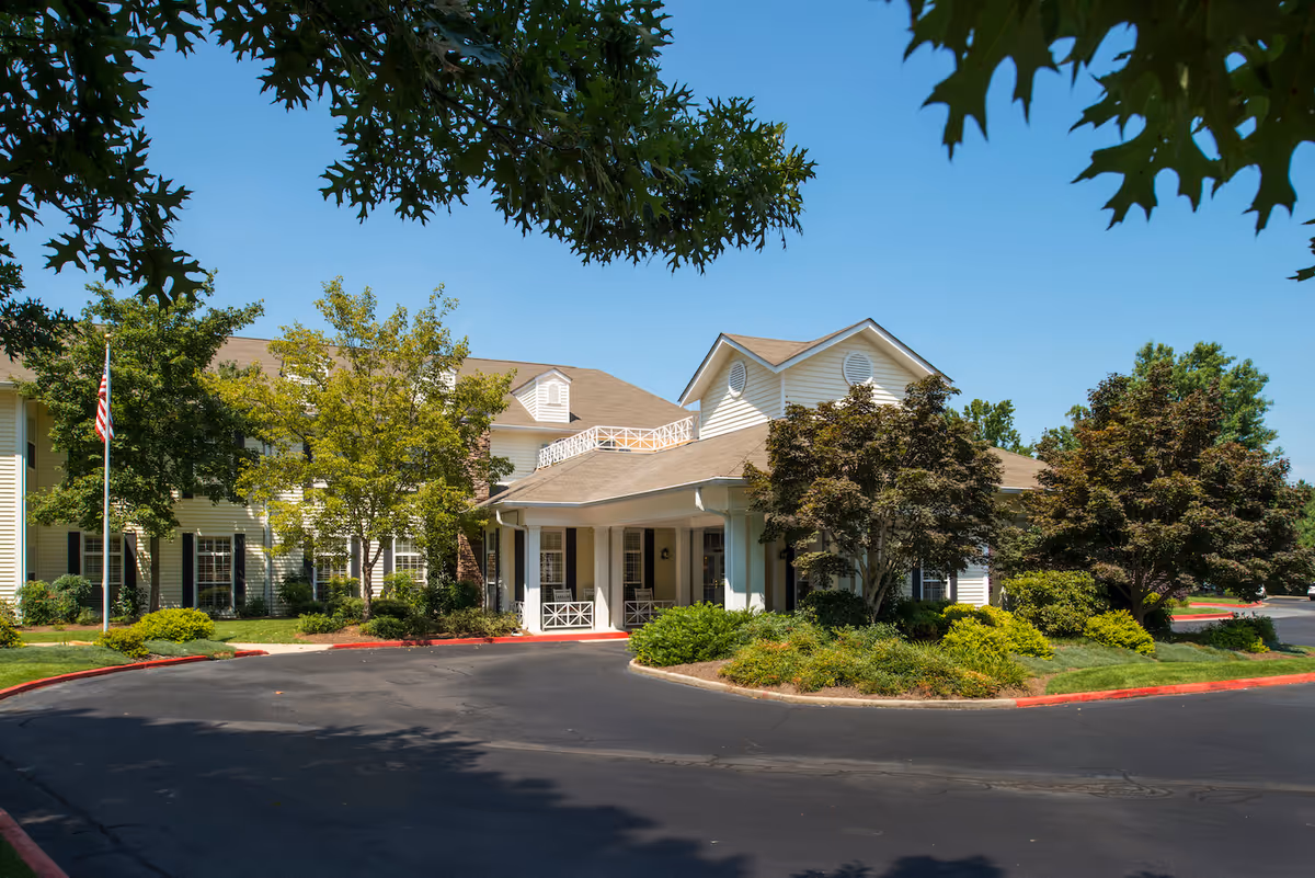 Exterior view of Arbor Terrace Athens senior living facility showing a large building with beige siding, multiple windows, a covered entrance with white columns, surrounded by green trees and shrubs under a clear blue sky.