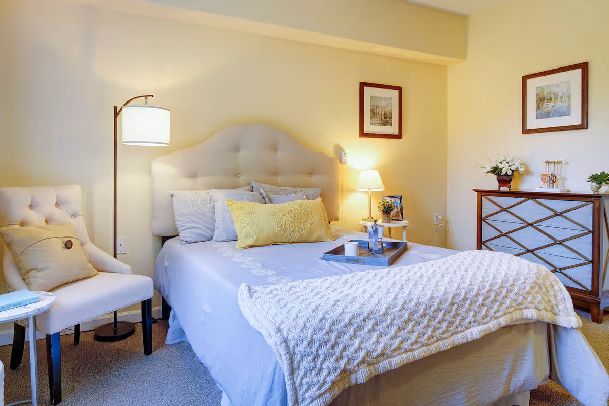 A cozy bedroom with a neatly made bed featuring a tufted headboard, multiple pillows including a yellow decorative one, and a textured white blanket. Next to the bed is a small round side table with a lamp and a tray holding a coffee pot and cup. A cushioned chair with a pillow is positioned beside a floor lamp. On the opposite side, there is a wooden dresser with decorative items and framed artwork hanging on the walls.
