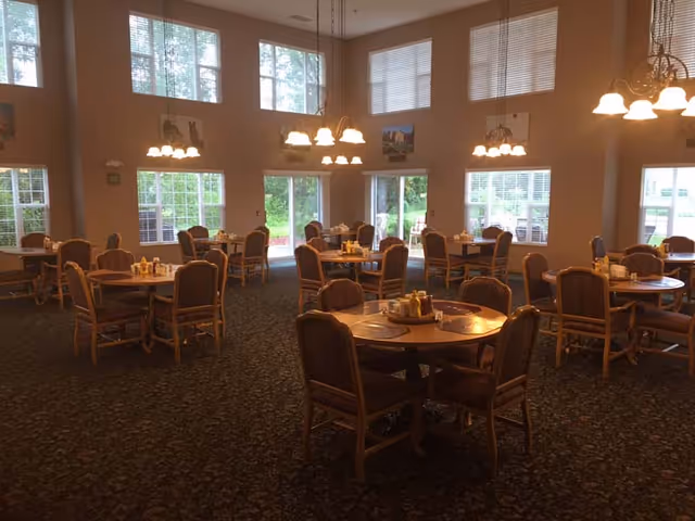 A spacious dining room with multiple round wooden tables, each surrounded by four cushioned chairs. The tables are set with placemats and condiment holders. Large windows let in natural light, and several hanging light fixtures illuminate the room. The walls are decorated with framed pictures, and the carpet has a dark patterned design.