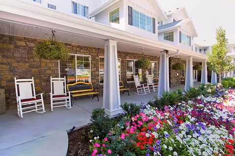 Front porch area of a senior living facility with white rocking chairs and benches under a covered walkway. The building has stone walls and white pillars, with hanging plants and colorful flower beds in the foreground. A person is sitting on the far right side near the flowers.