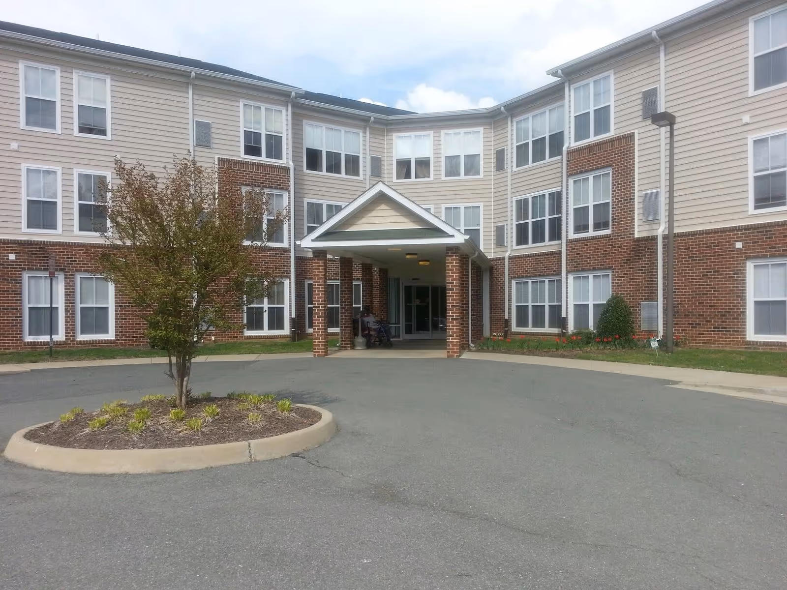 Exterior view of Sandston Plateau Senior Apartments showing a three-story building with a brick and beige siding facade. The entrance has a covered portico supported by brick columns. There is a small landscaped island with a tree and plants in front of the driveway leading to the entrance.