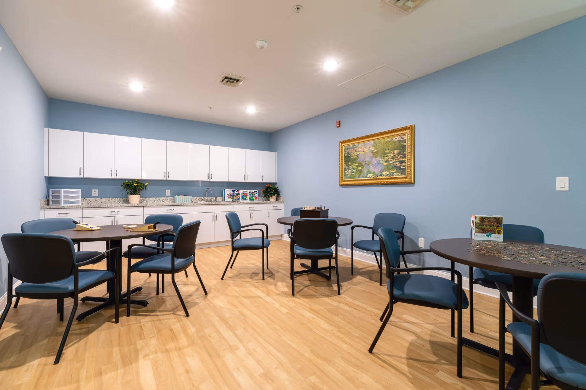 Well-lit communal activity/dining room with round tables and blue chairs facing a kitchenette with white cabinets against a light blue wall.