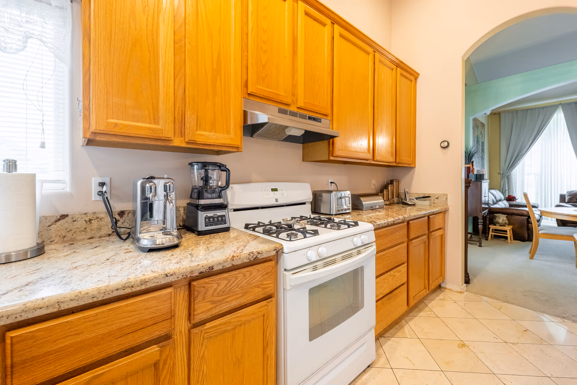 A kitchen with wooden cabinets, a white gas stove, a blender, a toaster, a coffee machine, and a paper towel holder on a granite countertop. The kitchen opens into a living room area with a dining table and chairs, and large windows with sheer curtains.