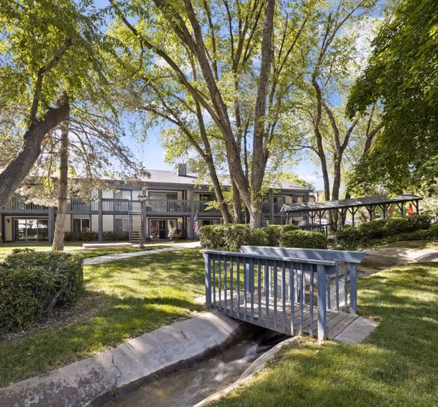 Outdoor view of a senior living facility with a small wooden bridge over a narrow stream, surrounded by green grass, trees, and bushes. The building in the background has two stories with balconies and a covered seating area to the right.