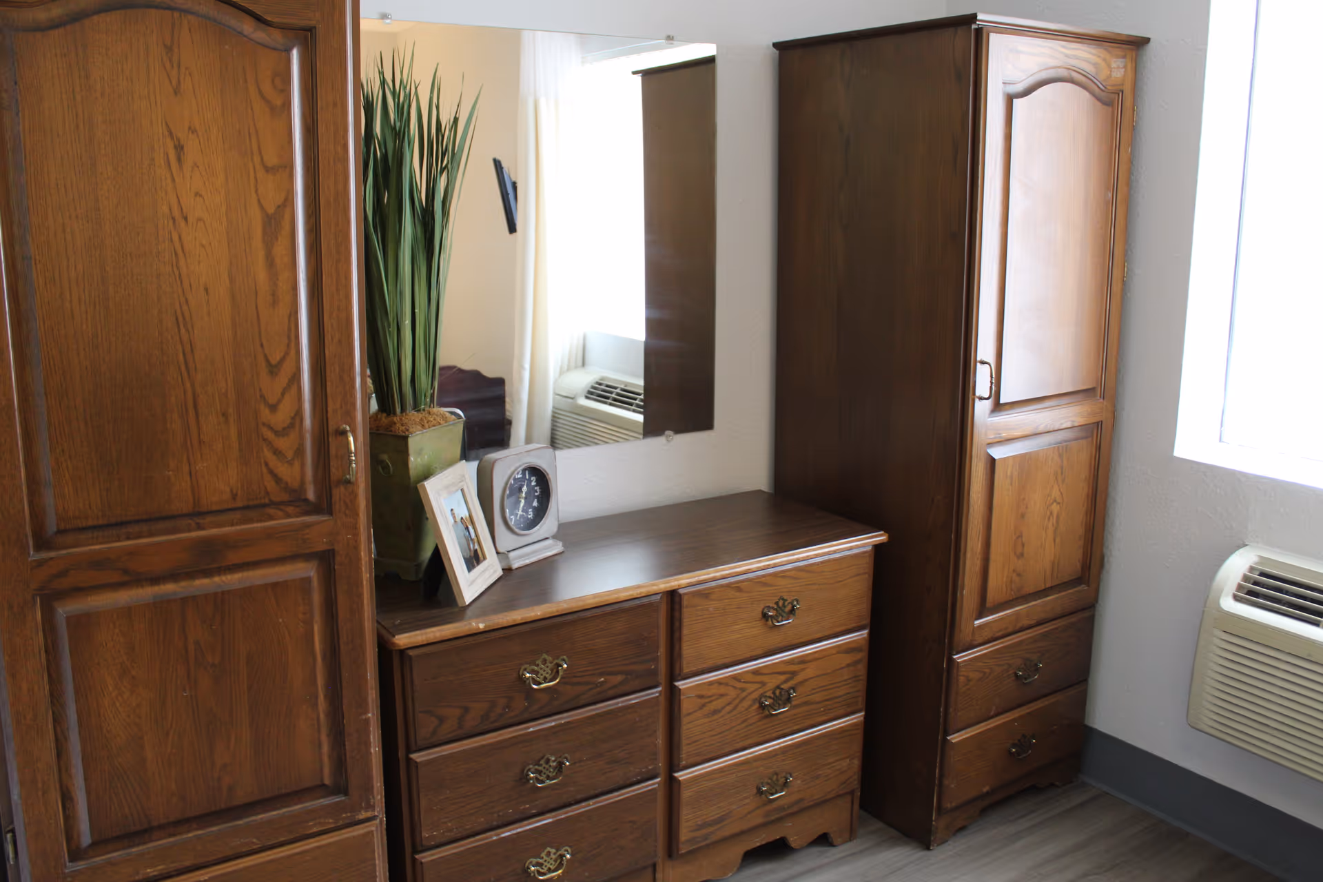 A room corner featuring wooden furniture including a tall wardrobe, a dresser with six drawers, and another tall cabinet. On top of the dresser are a potted plant, a framed photo, and a small clock. A mirror hangs on the wall above the dresser, reflecting part of the room with a window and an air conditioning unit.