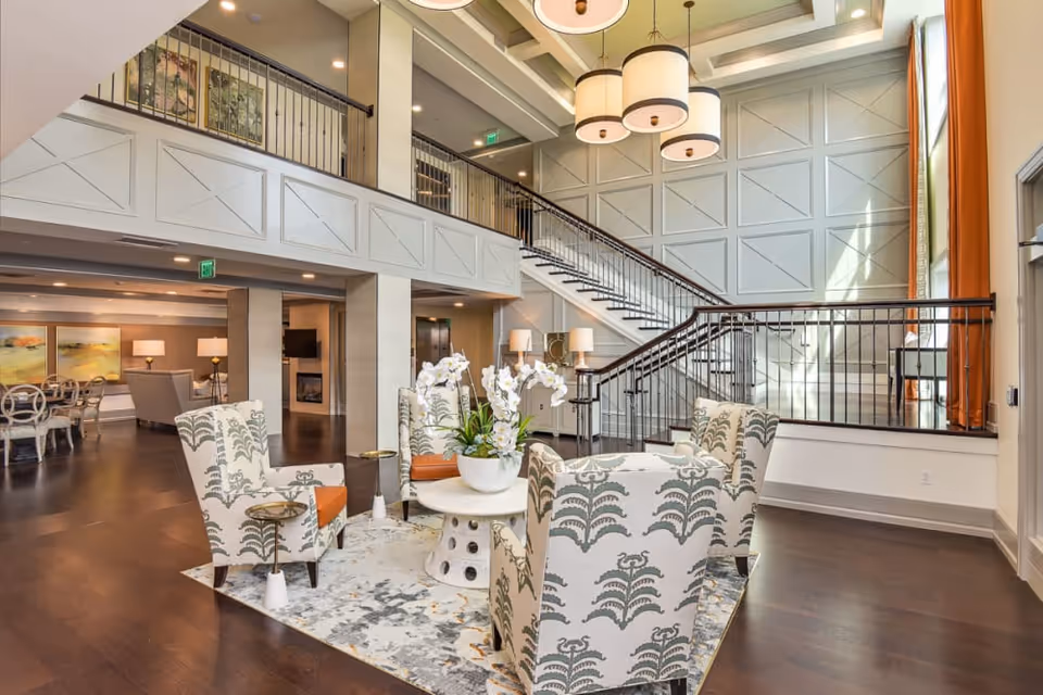 Spacious two-story senior living lobby with patterned armchairs around a central table, a grand staircase and suspended light fixtures.
