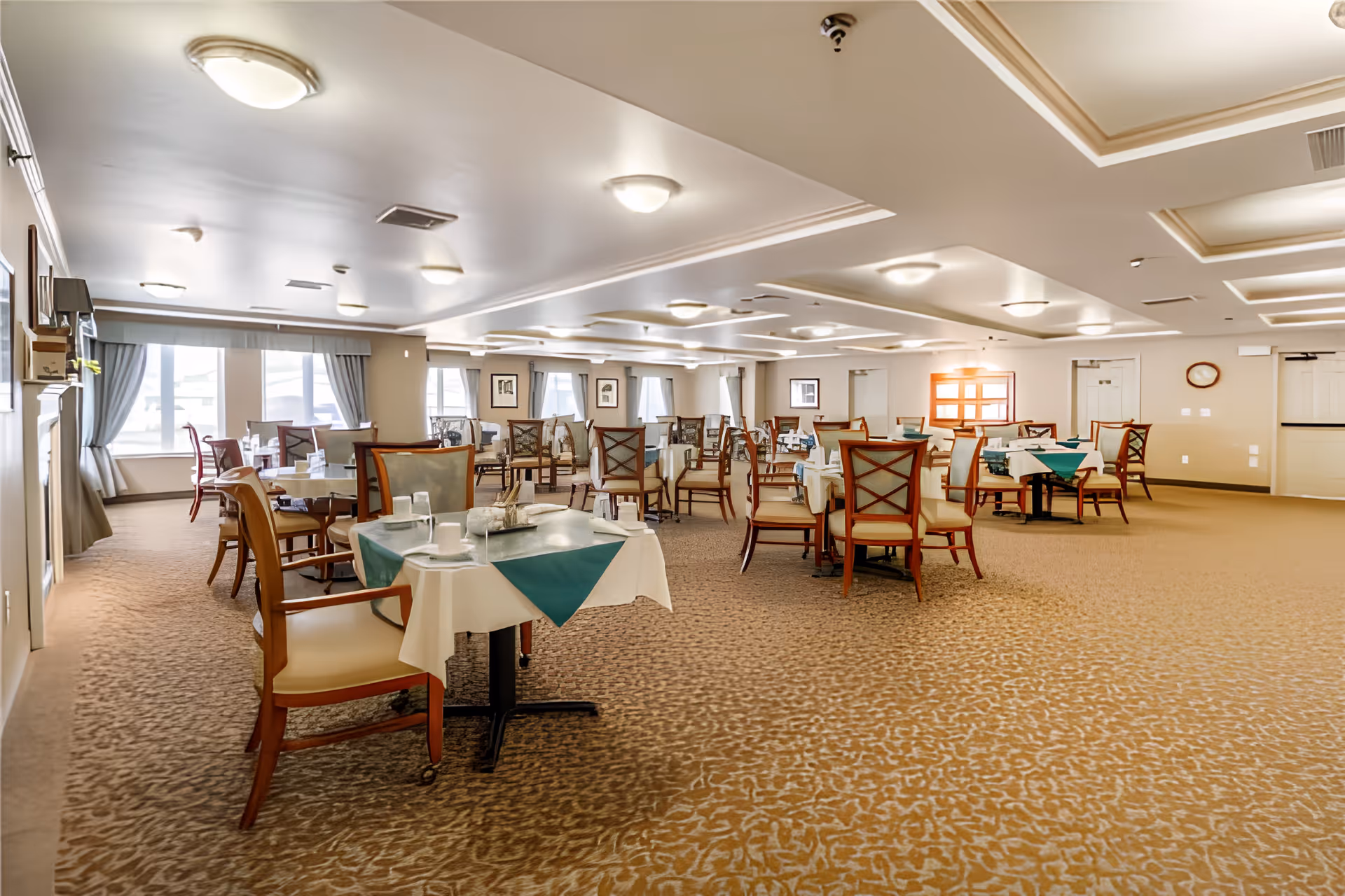 Spacious dining room with multiple set tables and wooden chairs, carpeted floor, and large windows with curtains.