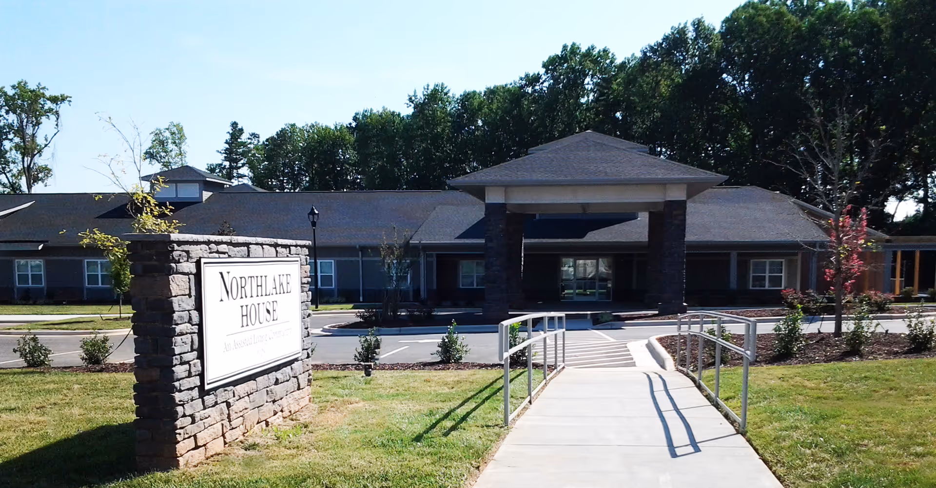 Exterior view of Northlake House, a single-story building with a covered entrance supported by stone pillars. A stone sign with the facility name is visible on the left side of a paved walkway leading to the entrance. The area is landscaped with grass, small bushes, and trees, with a background of taller trees under a clear blue sky.