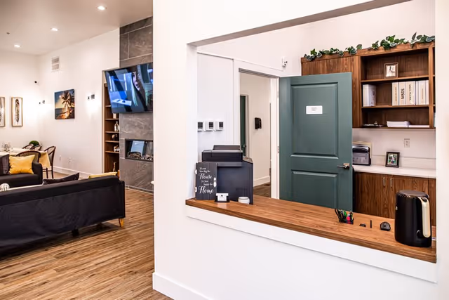 Interior view of a senior living facility showing a reception window with a wooden counter, office supplies, and a green door labeled 'Staff Only' behind it. To the left, there is a living area with black sofas, a dining table with chairs, framed artwork on the walls, a wall-mounted TV, and a fireplace with built-in shelves.