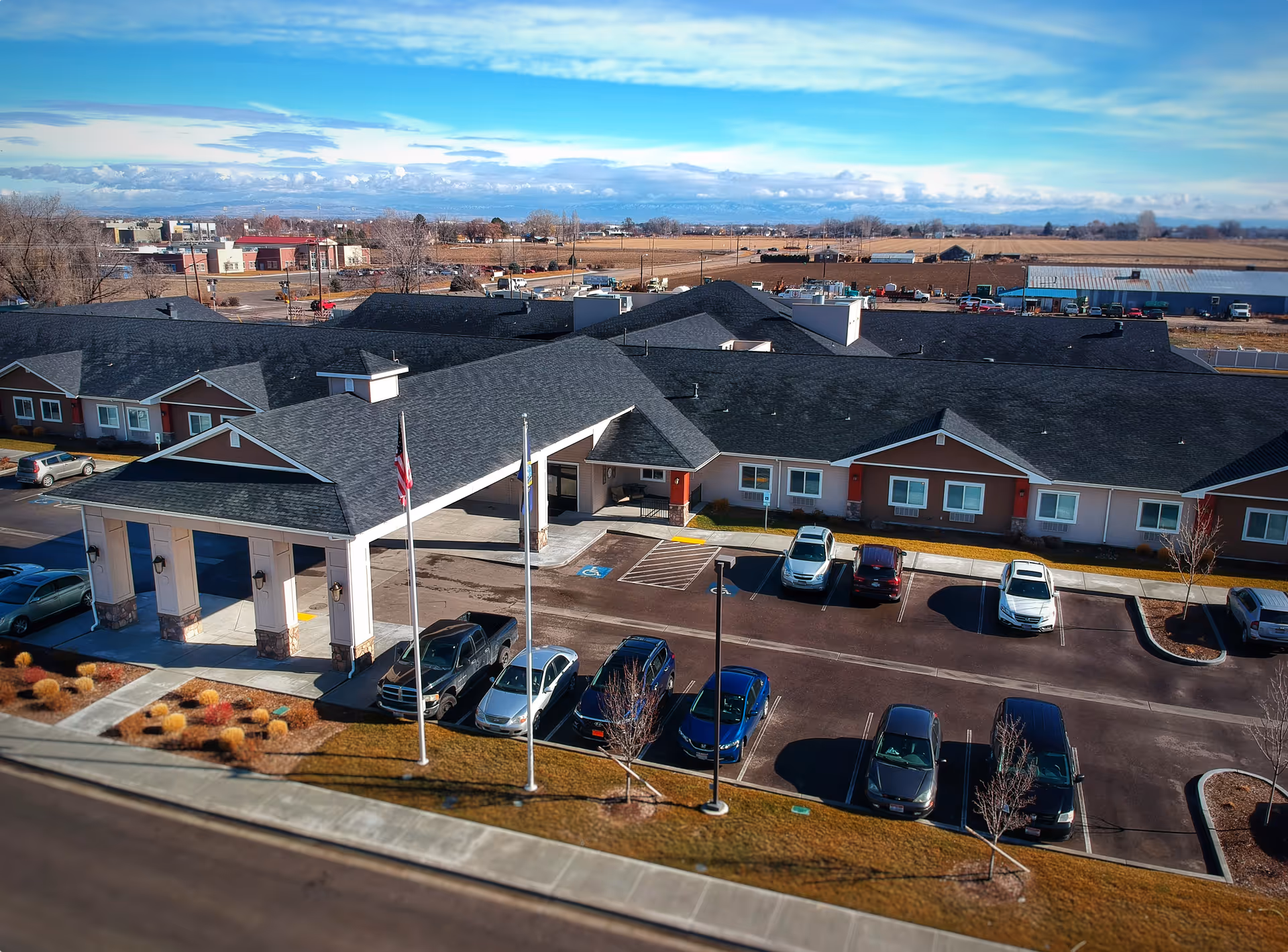 Aerial view of Aspen Creek Senior Living facility showing a single-story building with a covered entrance, multiple parked cars in the parking lot, and a surrounding landscape with trees and shrubs under a partly cloudy sky.