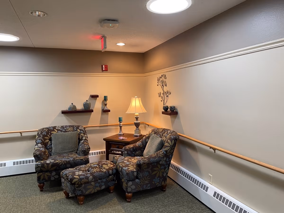 A cozy corner seating area in a senior living facility with two floral patterned armchairs, a matching ottoman, a wooden side table with a lamp and decorative candle holders, and wall-mounted shelves with decorative items. The walls are beige with a wooden handrail running along them.