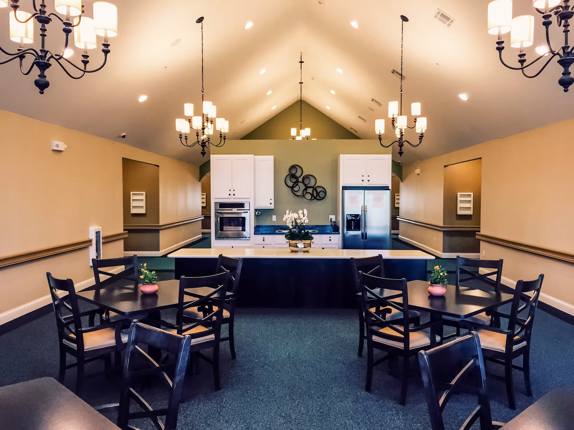 Interior view of a dining area with two square tables each surrounded by four dark wooden chairs. In the background, there is a kitchen area with white cabinets, a built-in oven, a refrigerator, and a central island with a flower arrangement. The room has a high vaulted ceiling with multiple chandeliers providing warm lighting.