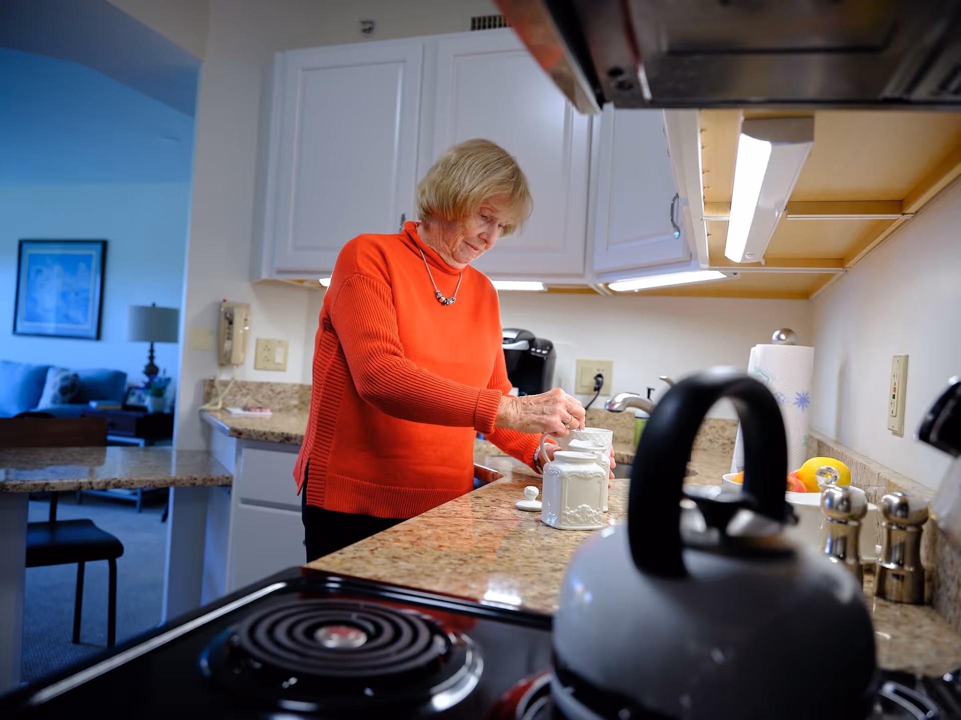 An elderly woman wearing a red sweater is standing in a kitchen, preparing something with white ceramic containers on a granite countertop. The kitchen has white cabinets and under-cabinet lighting. In the foreground, a stovetop with a kettle is visible. In the background, part of a living room with a blue couch and a framed picture on the wall can be seen.