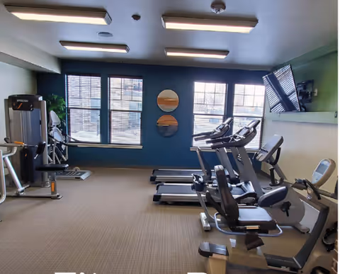 Interior view of a fitness room with exercise equipment including treadmills, stationary bikes, and weight machines. The room has large windows with blinds, two round mirrors on the wall, and a wall-mounted television.