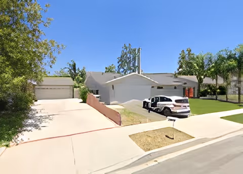 A single-story residential building with a driveway and two parked cars in front. The house has a white exterior with a garage and is surrounded by green trees and a well-maintained lawn under a clear blue sky.