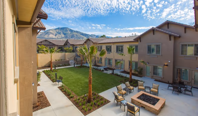 Sunlit courtyard with a central lawn, palm trees, patio seating and a fire pit surrounded by two-story stucco buildings and mountains in the background.
