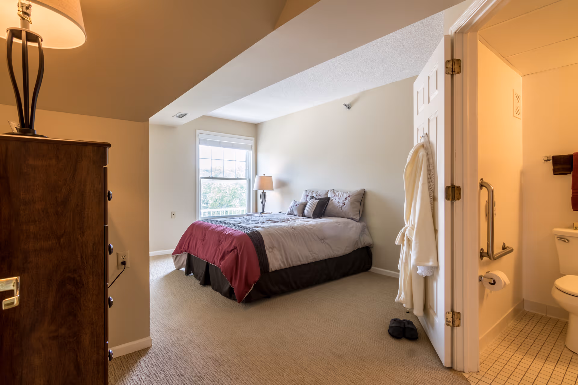 A cozy bedroom with a neatly made bed featuring gray and burgundy bedding, a window letting in natural light, a wooden dresser with a lamp on top, and an open door leading to a bathroom with a toilet and grab bars. A white robe and slippers are placed near the bathroom door.