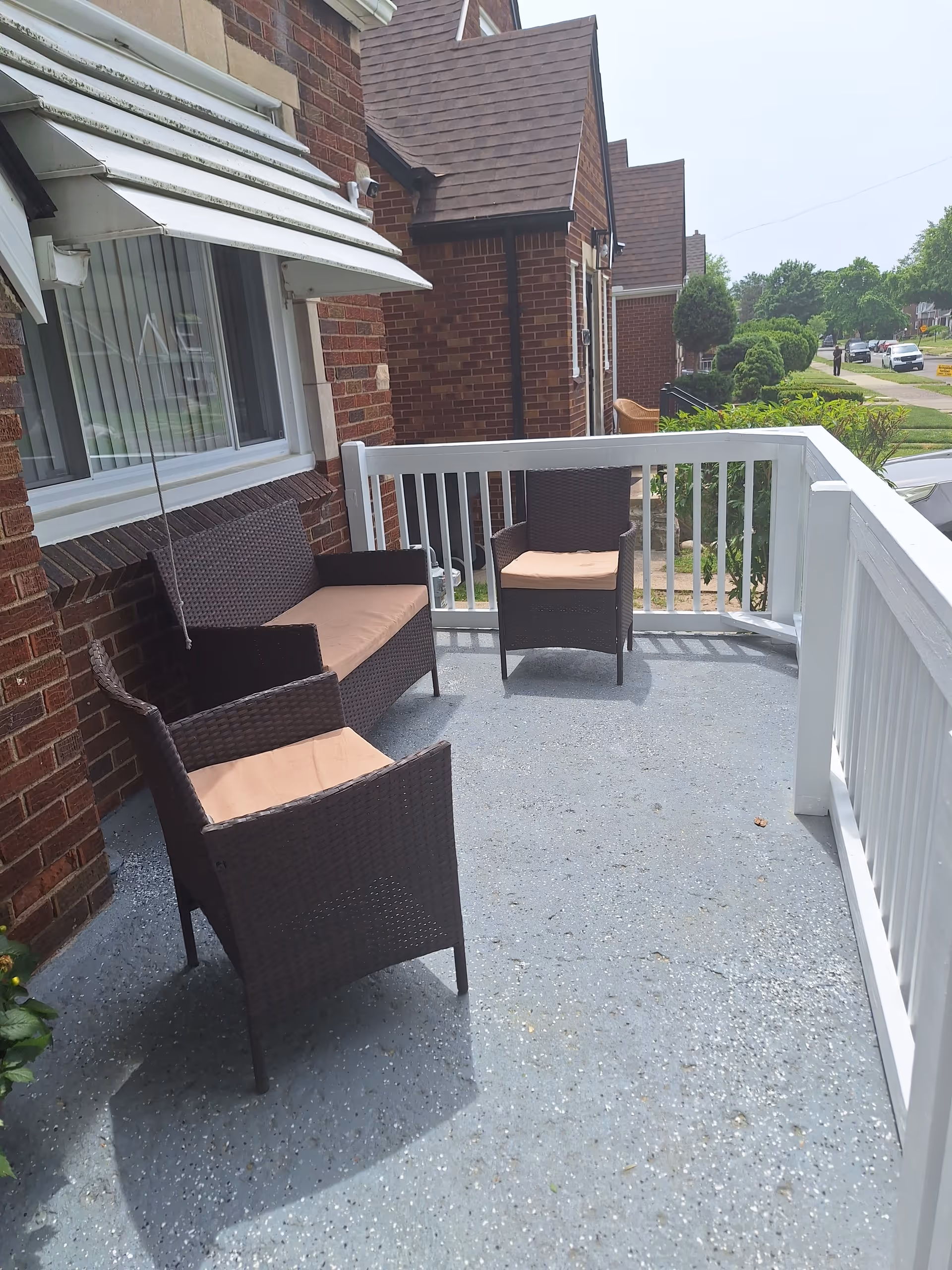 Front porch with wicker chairs and a bench on a painted concrete floor next to a brick house and white railing.