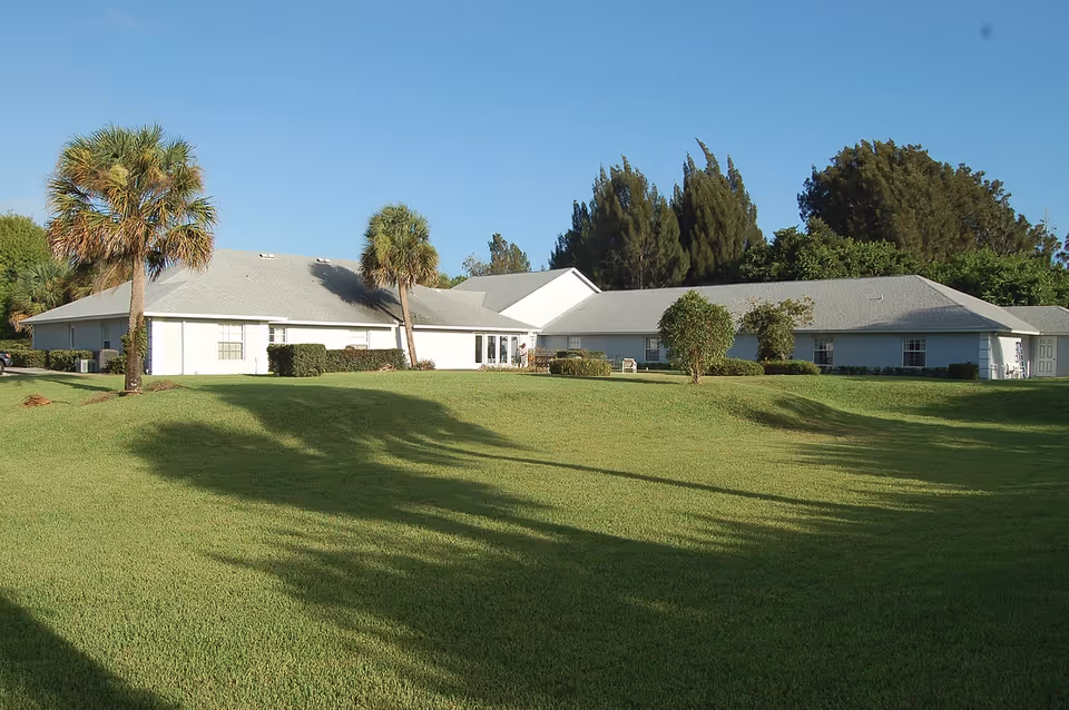 Single-story senior living facility building with a wide green lawn, palm trees, and a clear blue sky.