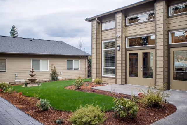 Exterior view of a senior living facility showing a landscaped courtyard with green grass, shrubs, and a multi-tiered water fountain. The building has beige siding, large windows, and glass doors leading to the courtyard.