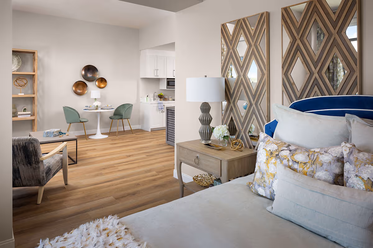 A bright and modern senior living apartment interior featuring a bed with multiple decorative pillows in the foreground, a wooden nightstand with a lamp and flowers, two large decorative mirrors on the wall, a cozy armchair, a small round dining table with two green chairs, and a kitchenette with white cabinets in the background. The room has wood flooring and neutral-colored walls.