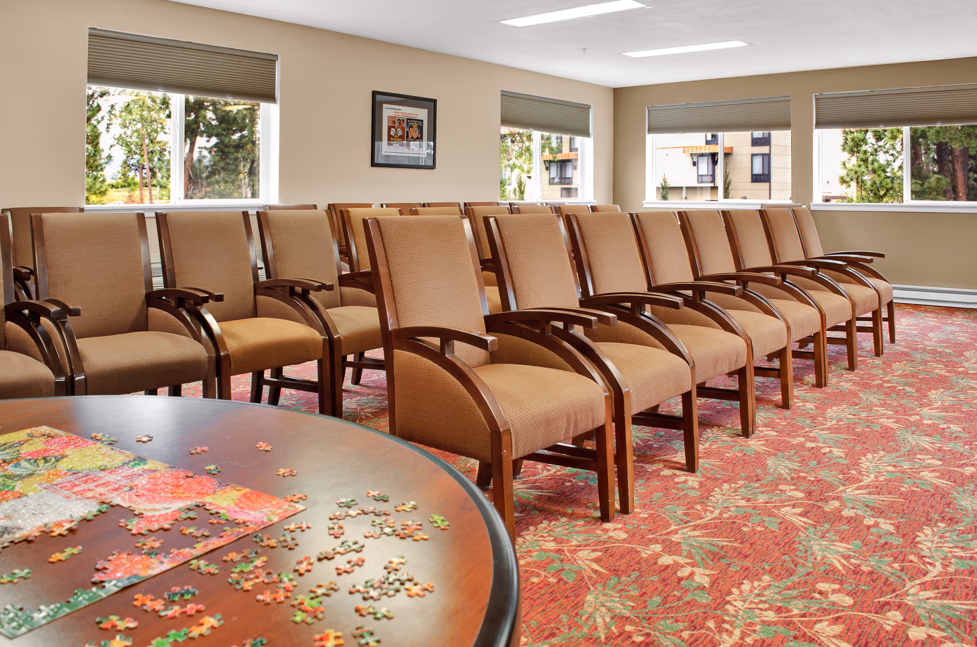 Rows of upholstered chairs in a communal activity room with a table in the foreground holding a partially completed jigsaw puzzle and windows along the far wall.