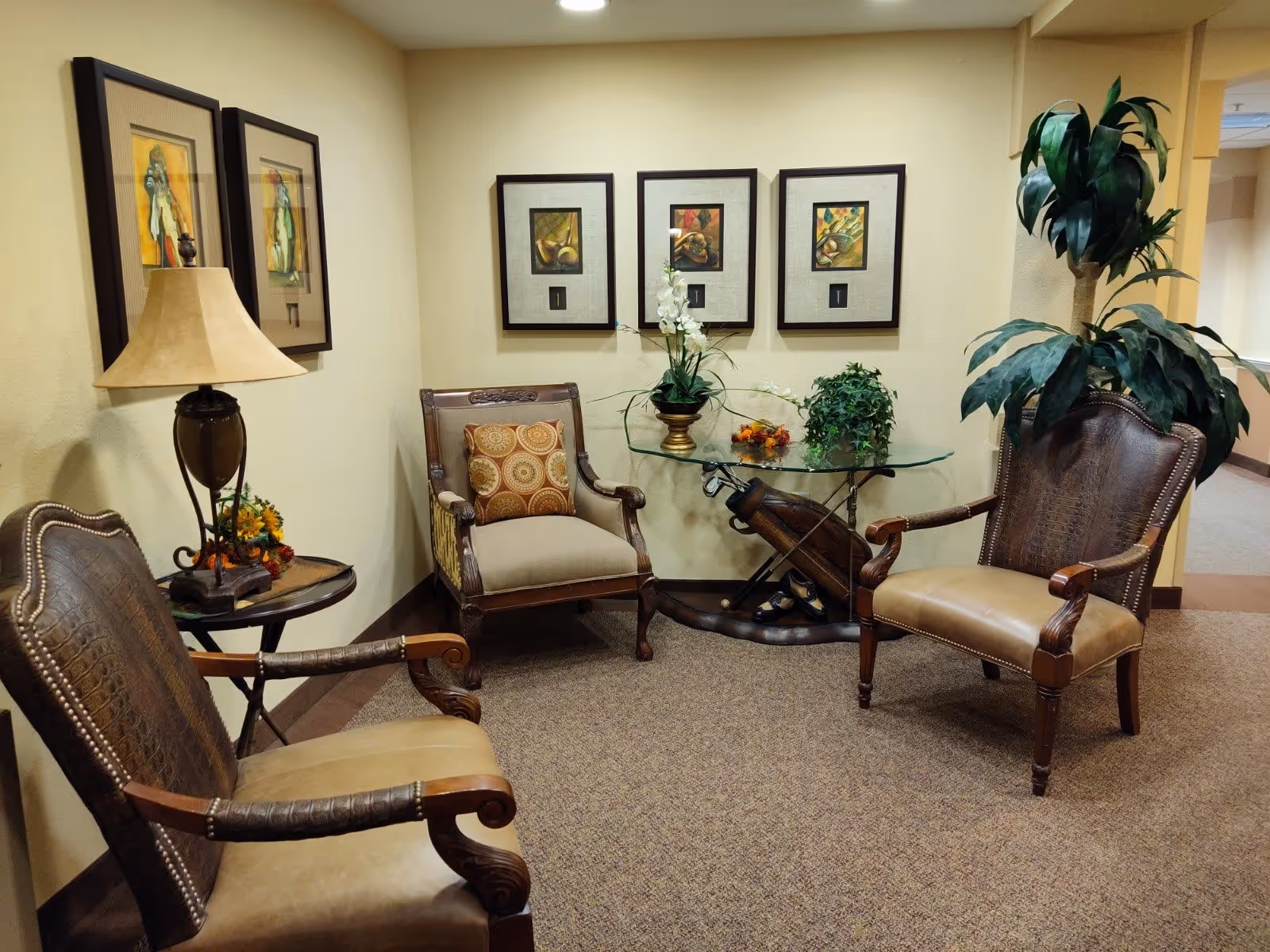 A cozy seating area in a senior living facility with three upholstered armchairs arranged around a glass-top table. The table has decorative plants and flowers on it. The walls are adorned with framed artwork, and there is a table lamp on a small side table next to one of the chairs. The carpeted floor and warm lighting create a welcoming atmosphere.