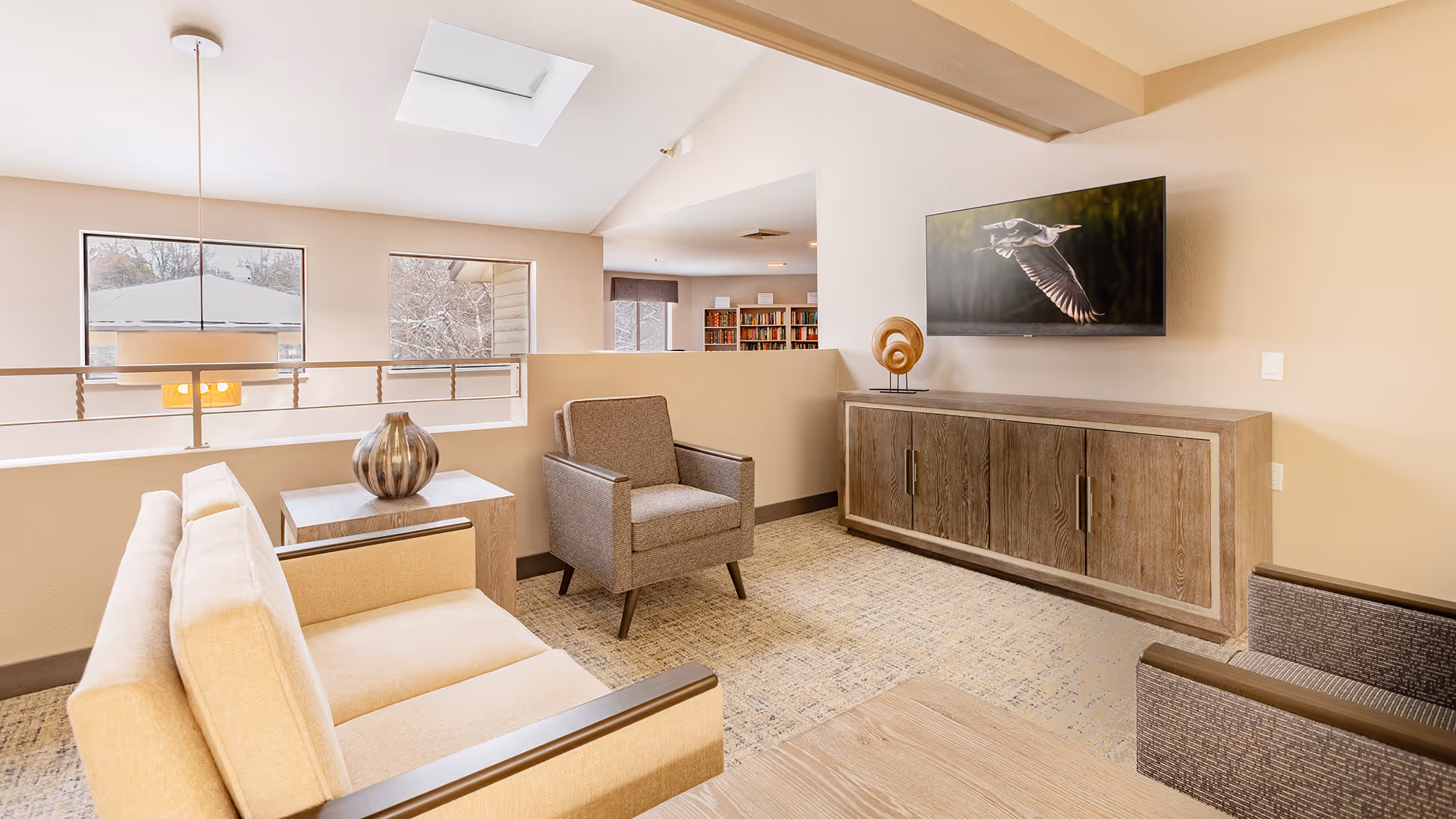 Bright lounge area with sofas and armchairs facing a wall-mounted TV above a wooden cabinet, with windows and a skylight.
