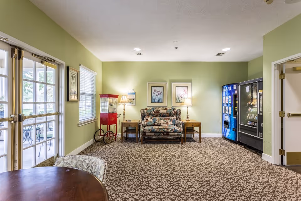 A common area in an assisted living facility with light green walls and patterned carpet. The room features a patterned loveseat between two wooden side tables with lamps, a red popcorn machine, and two vending machines. There is a round wooden table with a chair in the foreground and glass double doors leading to an outdoor patio with tables and chairs.