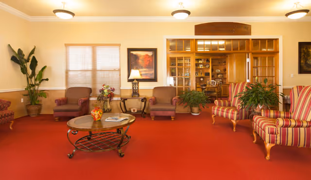A spacious lounge with red carpet, upholstered armchairs, a round coffee table and plants in a senior living facility.