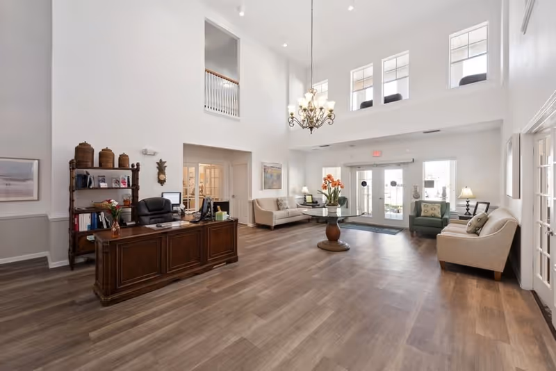 Bright, spacious senior living lobby with a wooden reception desk, seating area, chandelier, and double entrance doors.
