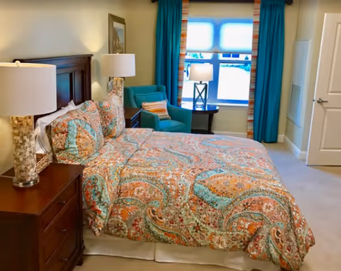 A cozy bedroom with a large bed covered in a colorful paisley-patterned bedspread. There are two bedside tables with matching lamps on either side of the bed. A teal armchair with a striped pillow is placed near a window with teal curtains and a striped valance. The room has beige walls and carpeted floor.