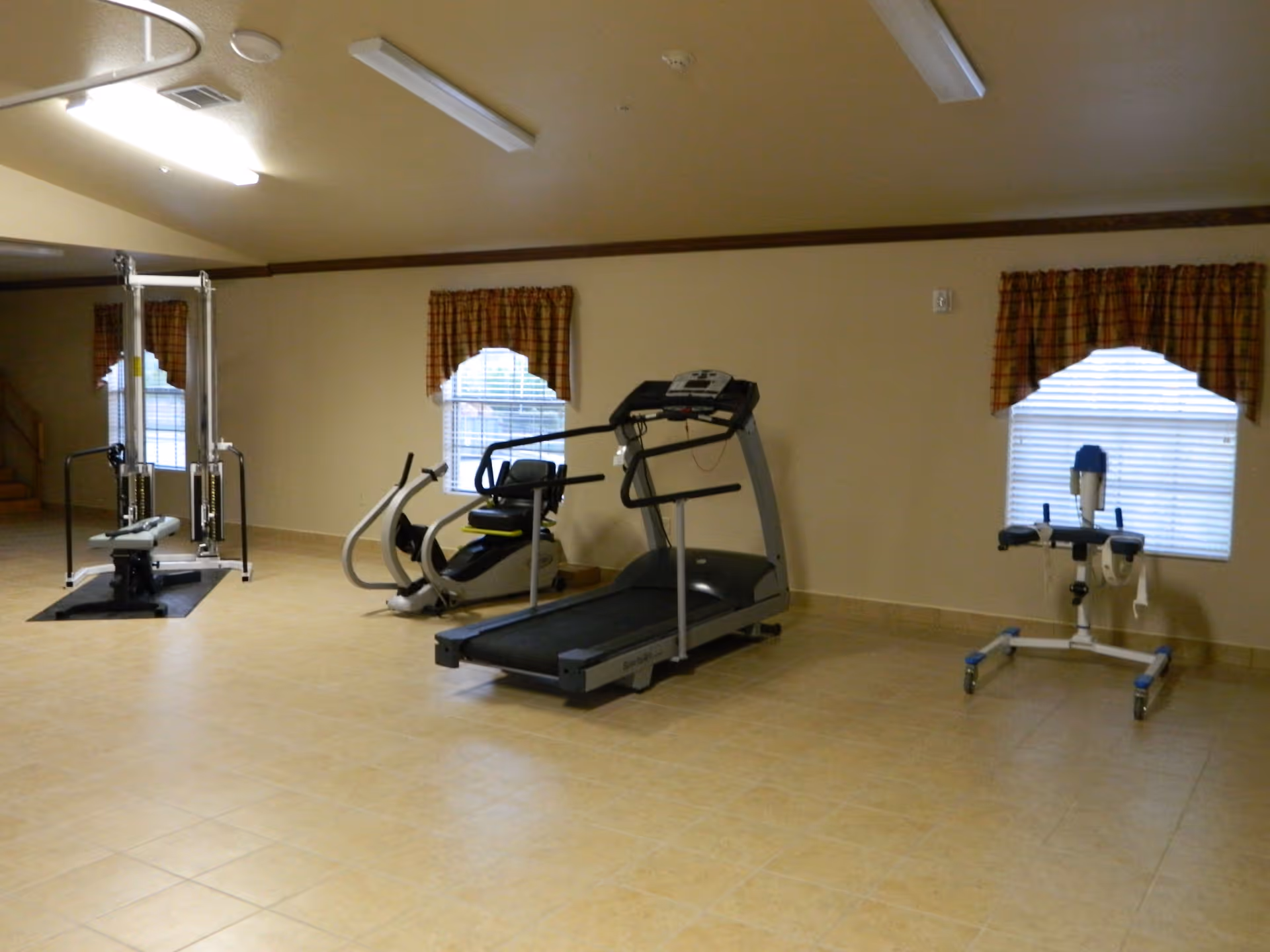 A fitness room with exercise equipment including a treadmill, a recumbent bike, a weight machine, and a standing exercise device. The room has tiled floors, beige walls, two windows with plaid curtains, and fluorescent ceiling lights.