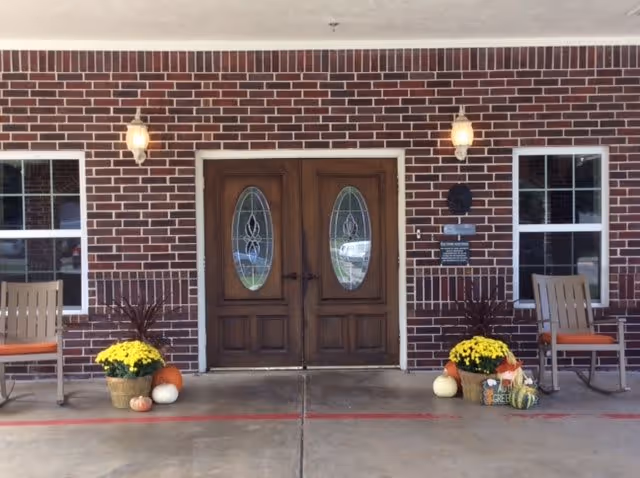 Front entrance of a brick senior living facility with double wooden doors, rocking chairs, and fall decorations including pumpkins and potted mums.