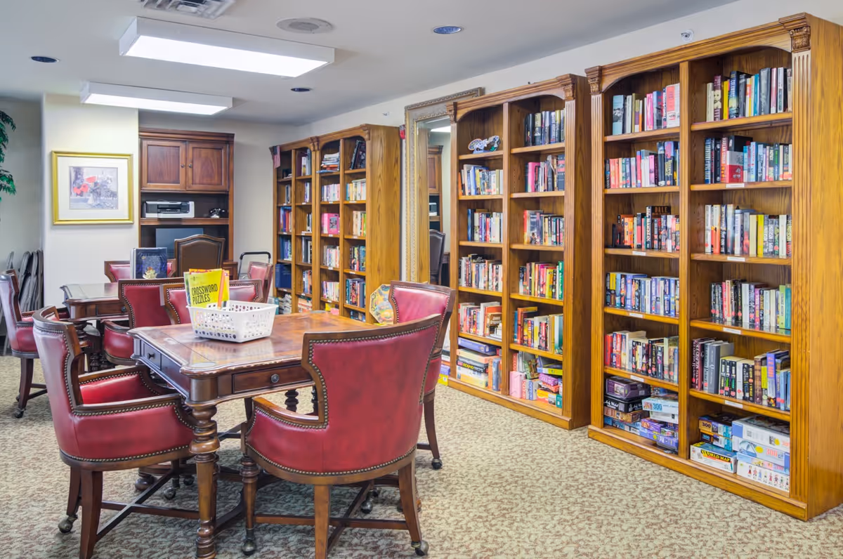A cozy library or reading room with wooden bookshelves filled with books and puzzles, a wooden table surrounded by red leather chairs, and a framed picture on the wall.