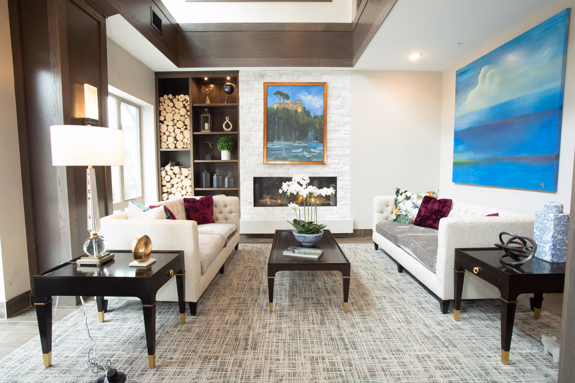 A bright and modern living room with two white tufted sofas facing each other, a dark wooden coffee table in the center with a potted orchid, and two matching side tables with decorative items and a lamp. The room features a white brick fireplace with a framed painting above it, built-in shelves with logs and decor, and a large blue abstract painting on the wall.