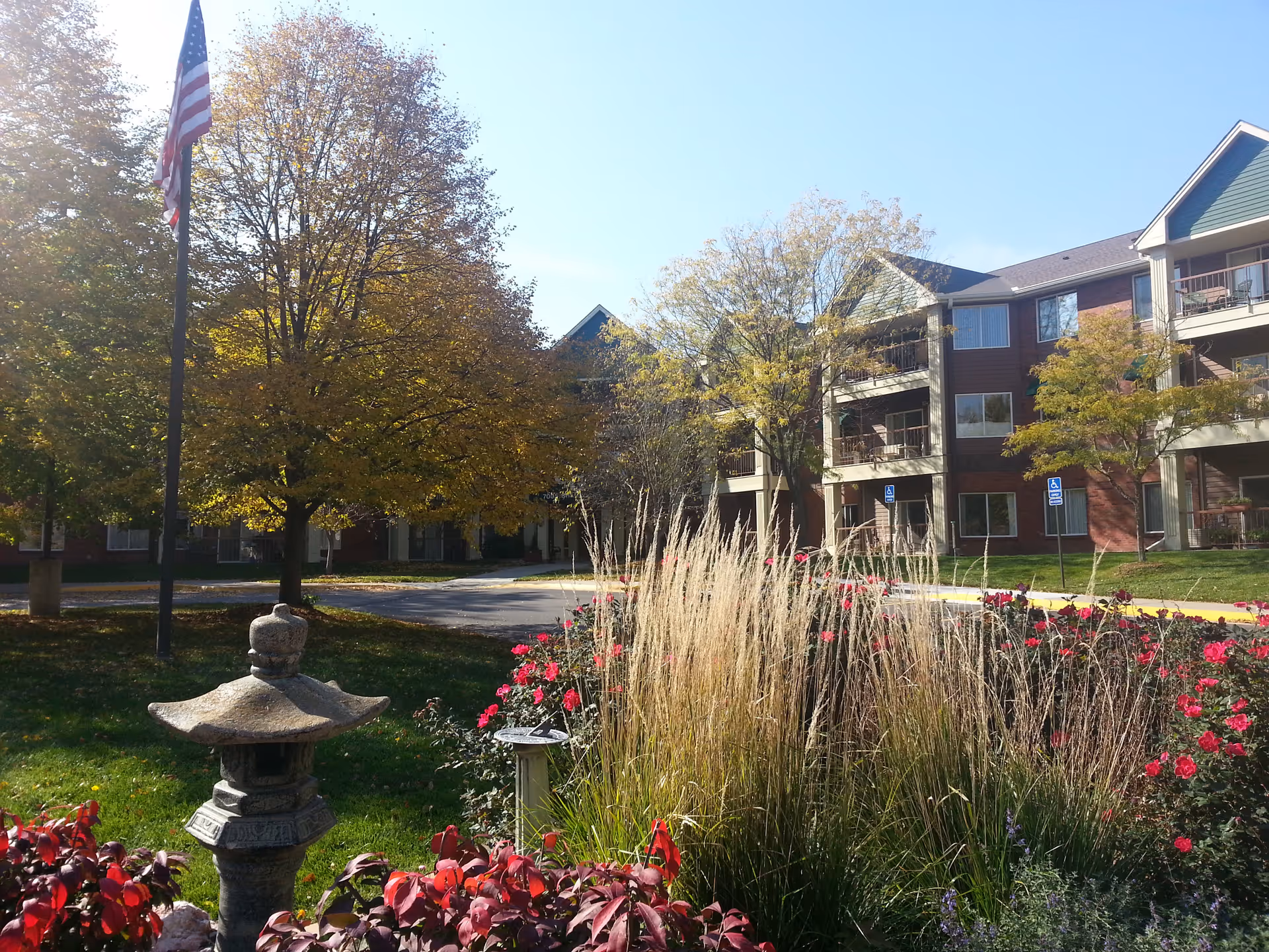 Outdoor view of a senior living facility named Carriage Glen with a landscaped garden featuring ornamental grasses, red flowers, and a stone lantern. The building is a multi-story brick structure with balconies, surrounded by trees with autumn foliage and a flagpole with an American flag.