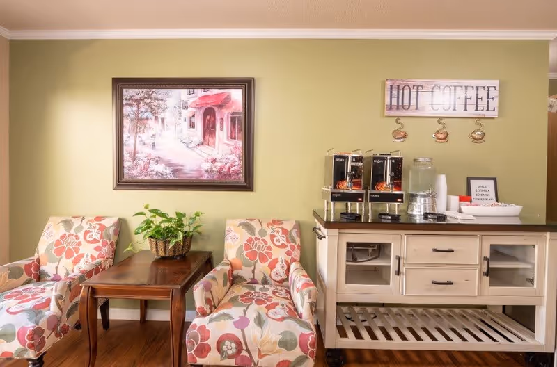 A cozy seating area with two floral-patterned armchairs separated by a wooden side table holding a green potted plant. On the right, a white cabinet with a dark countertop holds three coffee dispensers, a water dispenser, cups, and a small sign. Above the cabinet is a wall sign that reads 'HOT COFFEE' with decorative coffee cup hooks underneath. A framed painting of a street scene hangs on the light green wall behind the chairs.
