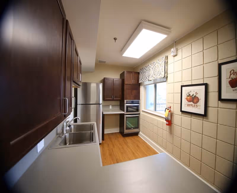 Small kitchen with wooden cabinets, a double sink, stainless steel refrigerator and oven, a window with a valance, and framed apple prints on a tiled wall.