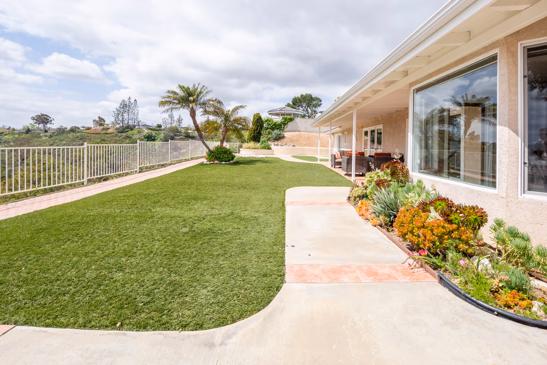 Outdoor area of a senior living facility with a well-maintained lawn, palm trees, a flower bed along the building, and a covered patio with seating. The area is fenced and overlooks a scenic landscape with hills and trees under a partly cloudy sky.