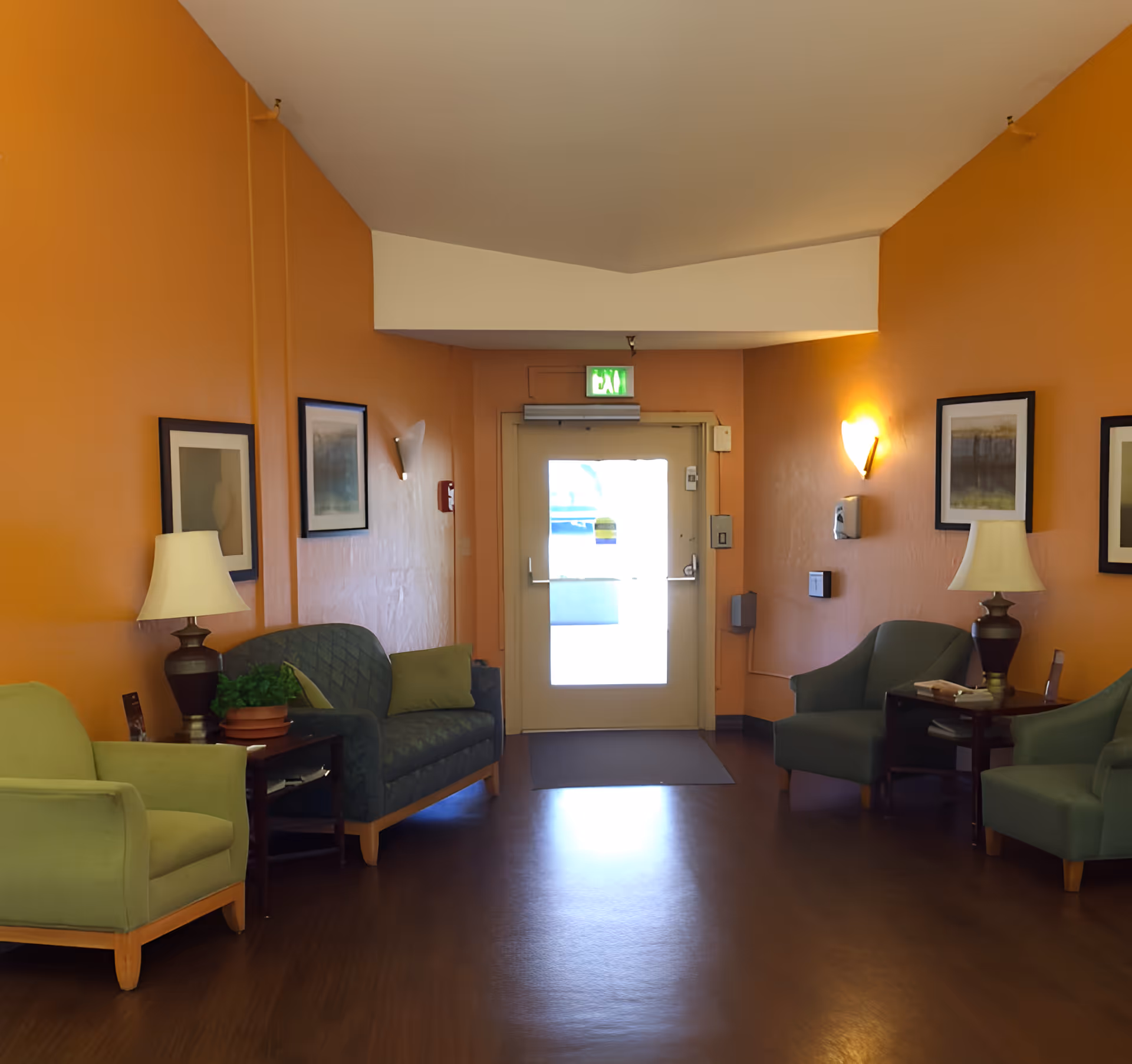 A cozy seating area in a hallway with orange walls, featuring two green armchairs and a green loveseat, two side tables with lamps and a potted plant, framed artwork on the walls, and a door with an exit sign above it at the end of the hallway.
