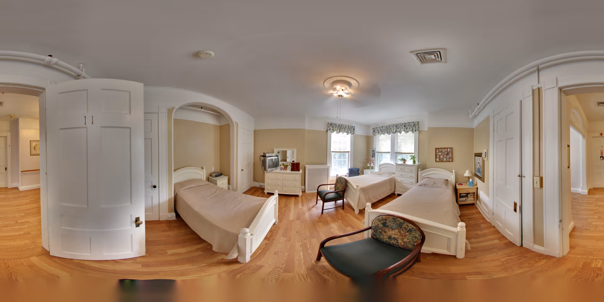 A spacious, well-lit bedroom in Riverview Lodge Residential Care Home featuring three single beds with beige bedspreads, wooden floors, two windows with floral valances, a dresser with a TV, and two armchairs with floral upholstery. The room has white doors and light beige walls, creating a warm and inviting atmosphere.
