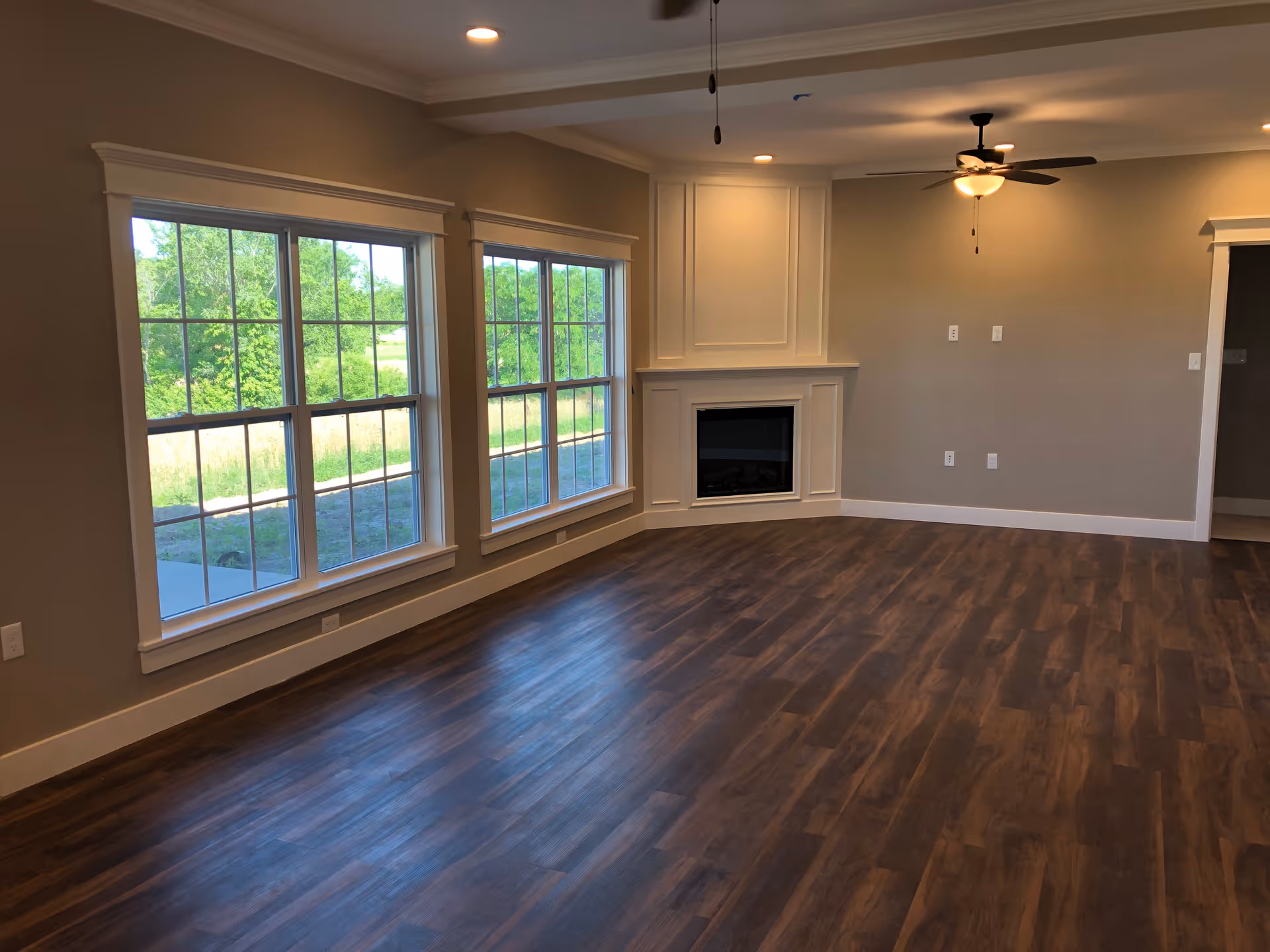 Empty living room with large windows showing greenery outside, a corner fireplace with decorative molding, ceiling fan with light, and wood flooring.