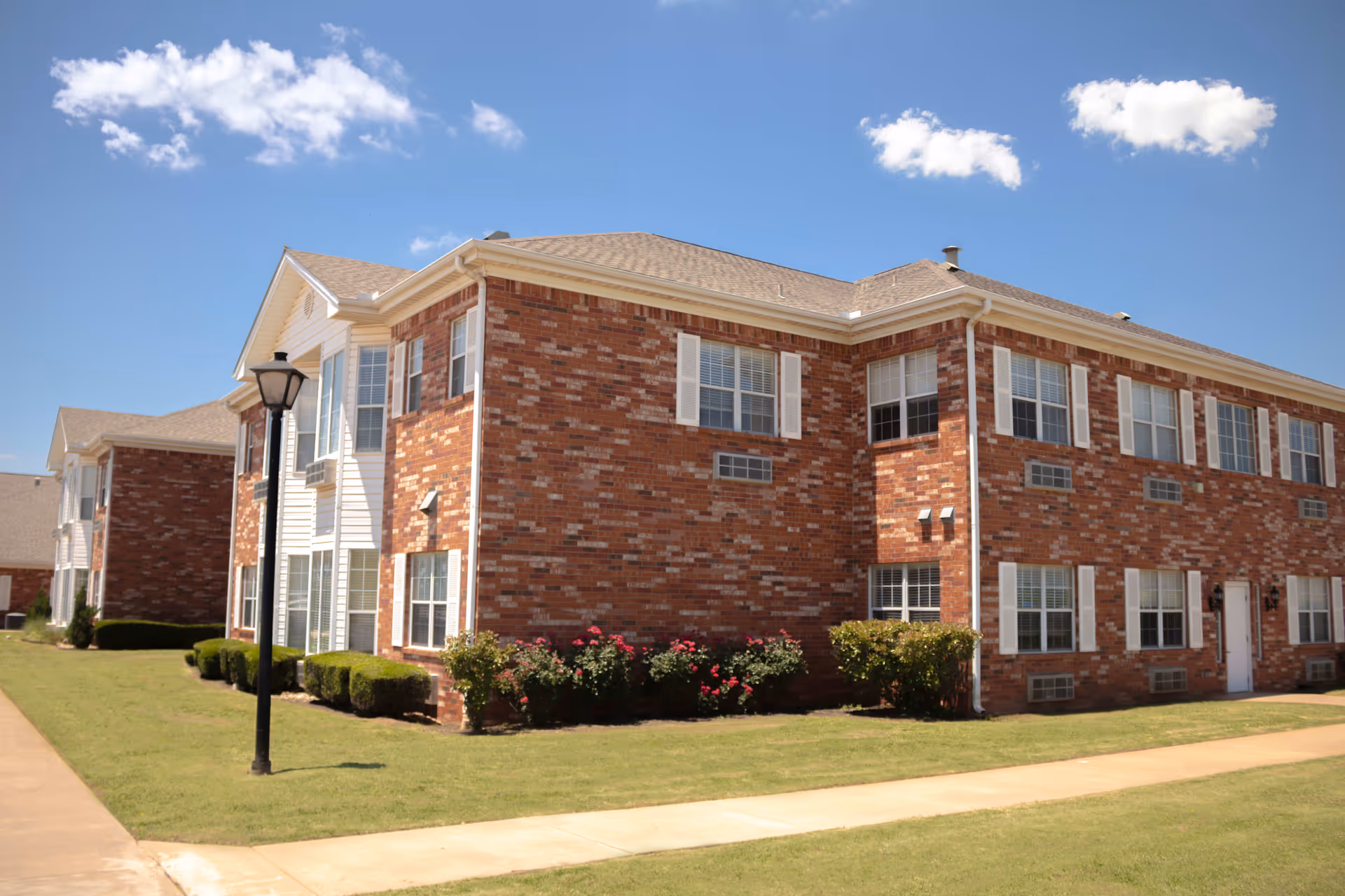 Exterior view of a two-story brick building with white window shutters and a beige roof under a blue sky with a few clouds. The building is surrounded by a well-maintained lawn, bushes, and a sidewalk with a black street lamp.