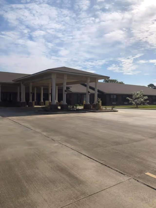 Exterior view of a single-story nursing and rehabilitation facility with a covered entrance supported by columns, a large concrete driveway, and a partly cloudy sky.