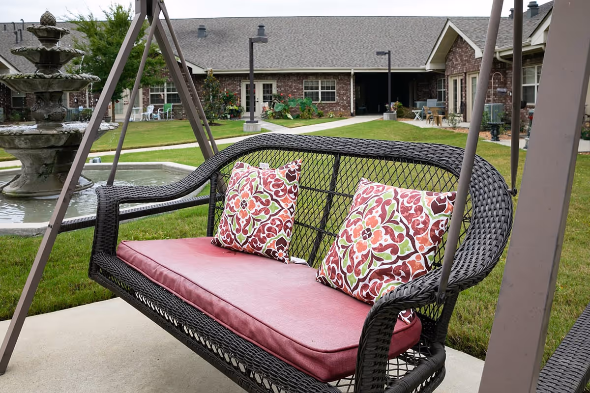 Outdoor swing bench with a maroon cushion and two decorative pillows with red, green, and white patterns, set on a concrete patio. In the background, there is a multi-tiered water fountain, green grass, pathways, and a brick building with windows and doors.